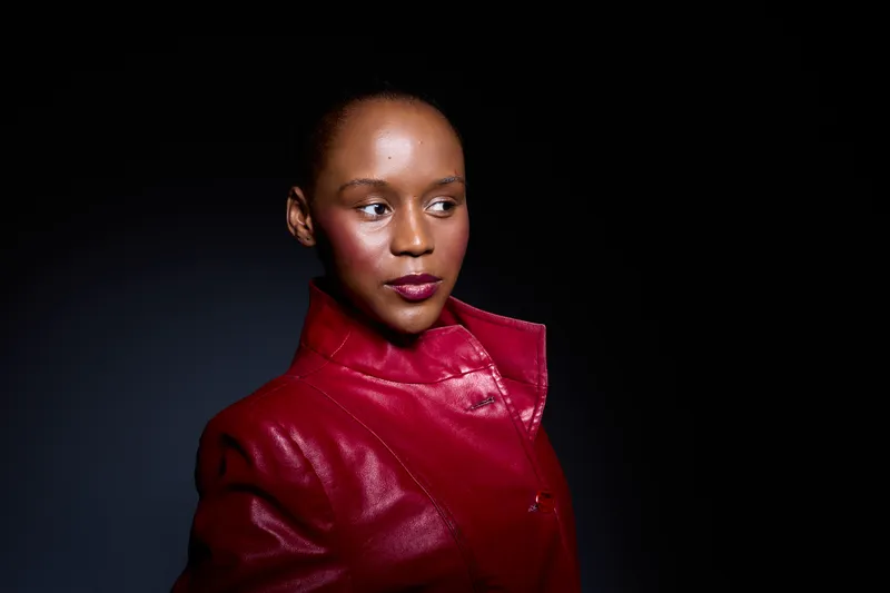 Portrait of confident black woman with dark skin wearing shiny red leather jacket. Studio lighting highlights her face and stylish clothing on black background. Fashion and beauty concept.
