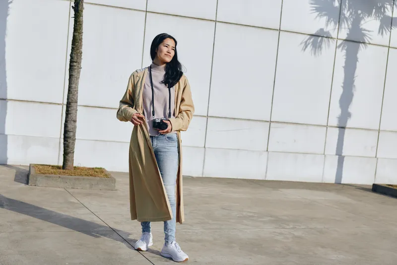 Woman in a long beige coat, light sweater and jeans sits outdoors near a white wall, holding a camera, relaxed pose, urban style, natural light, candid fashion moment