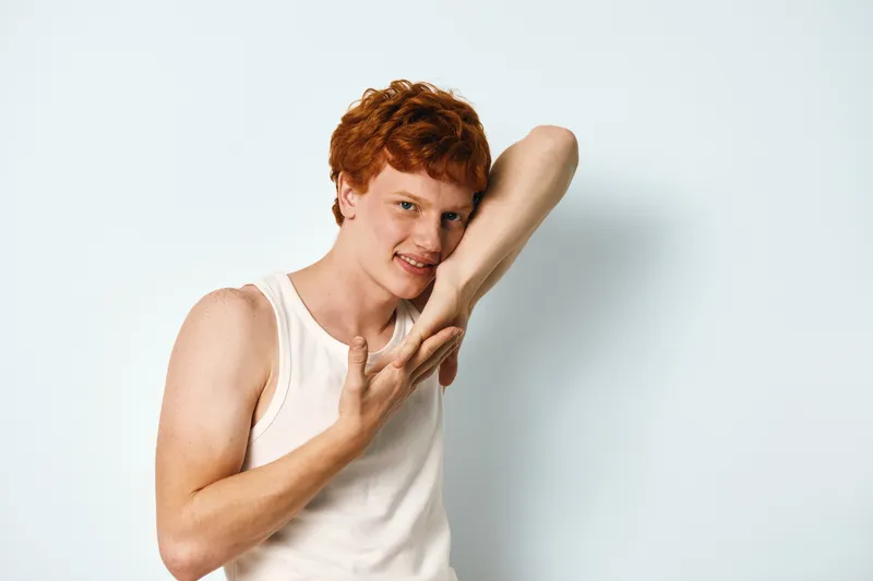 Redhead young man with freckles posing in sleeveless shirt on white background, casual expression and relaxed posture in closeup portrait studio.