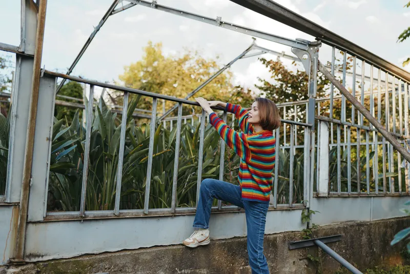 Woman climbing fence at a greenhouse among tall plants outdoors, wearing a colorful sweater and casual jeans and sneakers, gardening exploration and leisure in sunlit autumn backyard.