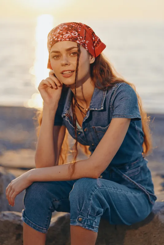 young woman wearing red bandana and denim outfit sitting by the sea during golden hour with relaxed and thoughtful expression