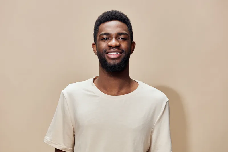 Smiling Black man in a casual beige t shirt, showcasing joy against a neutral background, emphasizing modern lifestyle and positivity