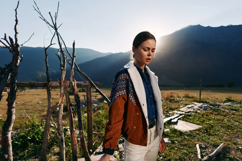 Woman in nature by mountain field at sunset, fashion portrait wearing textured jacket and shirt, outdoor lifestyle scene with rustic fence, contemplative model and dramatic backlight glow.