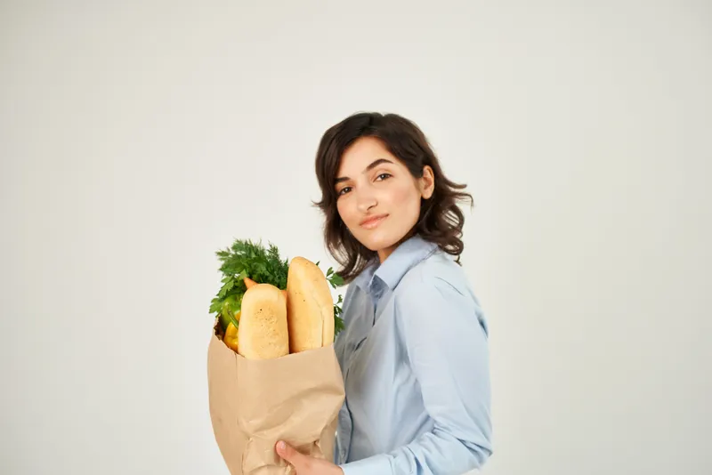 Симпатичная женщина с пакетом продуктов, овощами и здоровой пищей на светлом фоне