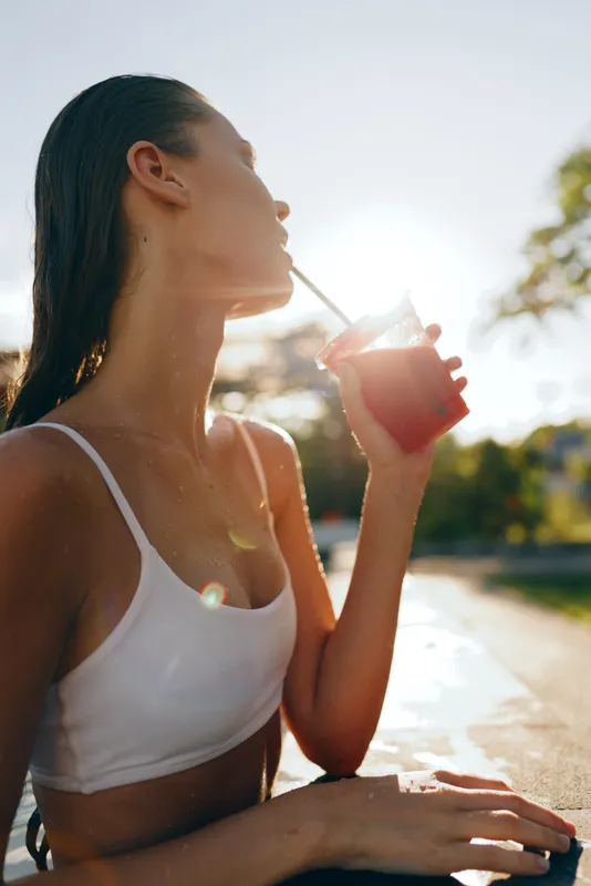 Young woman in white sportswear drinking juice from glass with straw outdoors. Sunny day creates warm natural light and healthy lifestyle concept in urban park setting.