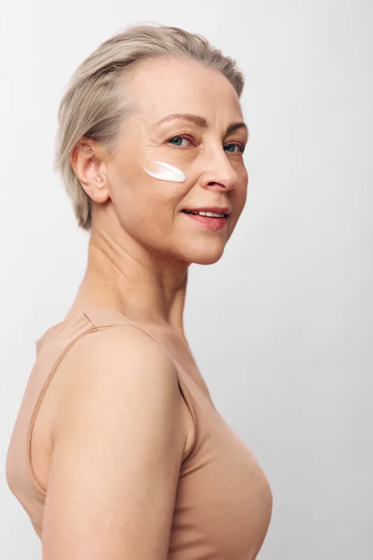 Middle aged woman with short grey hair applies face cream on cheek, wearing beige top against white background. Concept of skincare, anti aging, beauty routine, and natural wellness.