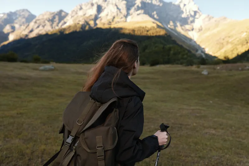 A lone hiker with a large backpack stands in a grassy alpine valley, gazing toward rugged mountains under a clear blue sky, capturing a dynamic outdoor adventure mood and vast landscape.