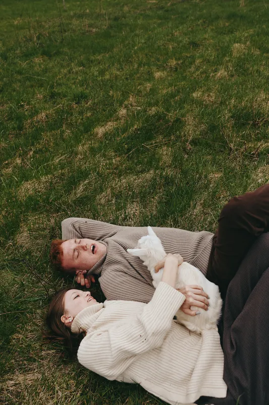 Relaxed couple lying on grass with a goat kid nearby, embracing each other in a cozy outdoor setting. Warm light highlights affection, nature, and simple lifestyle moments.