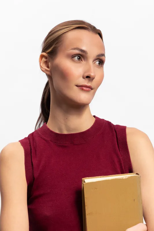 Young woman with tied hair wearing sleeveless burgundy top holds closed book, looking sideways on white background. Concept of education, knowledge, and thoughtful focus.