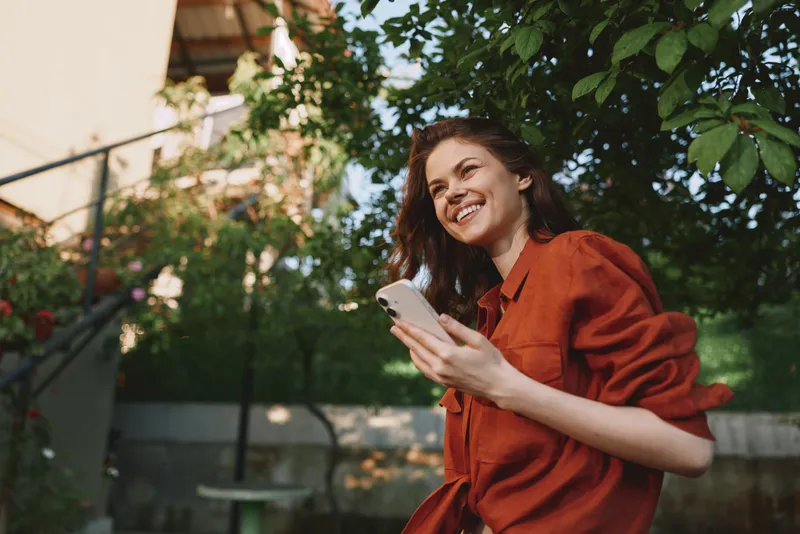 Vibrant outdoor moment with a smiling woman, casualwear, holding smartphone, sunlight and breeze, lush greenery in background, lifestyle focused portrait in natural colors