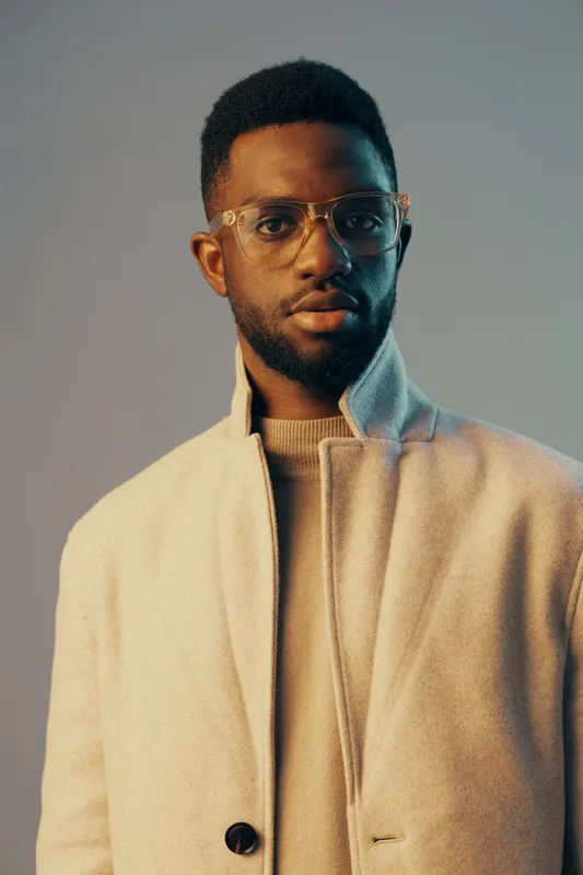 Stylish African man in a beige overcoat with glasses, showcasing confidence in a soft pastel background, ideal for fashion, portrait, and seasonal themes