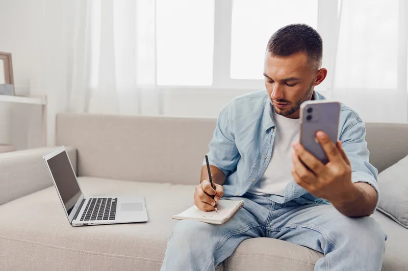 Focused man engaging with mobile device while jotting notes, showcasing productivity and modern work-life balance in a bright, cozy home office.