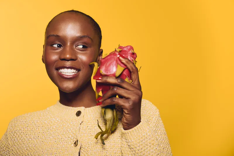Smiling young black woman with dark skin holds a dragon fruit near her face, wearing a beige sweater against a yellow background. Concept for healthy lifestyle and natural food.
