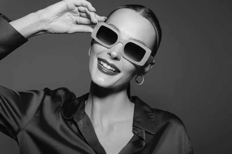 Smiling white European woman with fair skin wearing oversized sunglasses and silk shirt poses confidently against black background. Concept of fashion, style, and modern elegance.