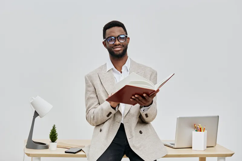 Professional smiling man with glasses, wearing beige blazer and white shirt, holding open book, standing in modern office with white background, desk, laptop, stationery, and small plant, conveying ed