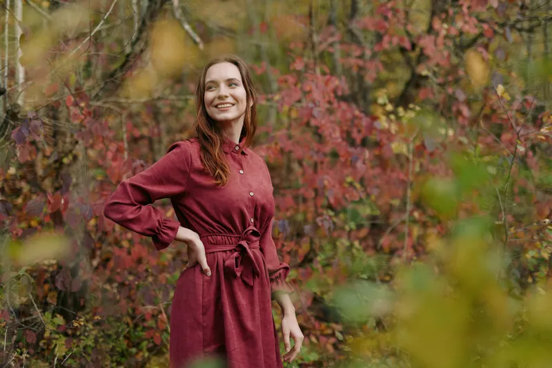 A candid portrait of a woman in a red dress, smiling softly, standing among autumn foliage. The scene conveys authentic mood and veracity through natural light and relaxed posture.