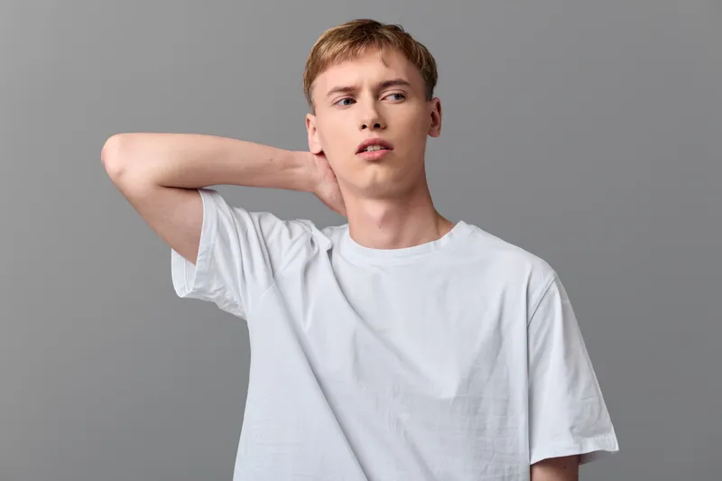 Young man in a plain white tee shows discomfort with a hand at the neck in a minimal studio. A casual lifestyle shot highlighting fashion, emotion and fatigue for contemporary market banners and