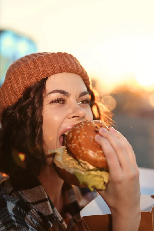 Woman eating burger outdoors at golden hour, candid lifestyle portrait with golden hour glow, authenticity and mindful living vibe, emotional storytelling in urban sunset.