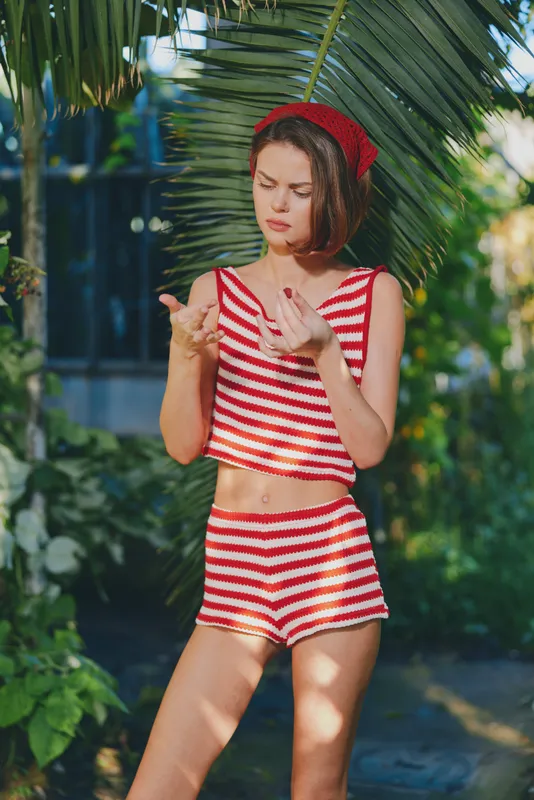 A cheerful young woman wearing a red and white striped crop top and shorts stands outdoors amid lush tropical plants, exuding playful summer energy and confident presence.