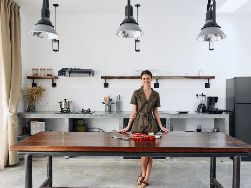 Счастливая женщина стоит в полный рост в стильном минималистичном интерьере кухни с натуральными элементами из дерева и камня, на столе свежие продукты для ужина
