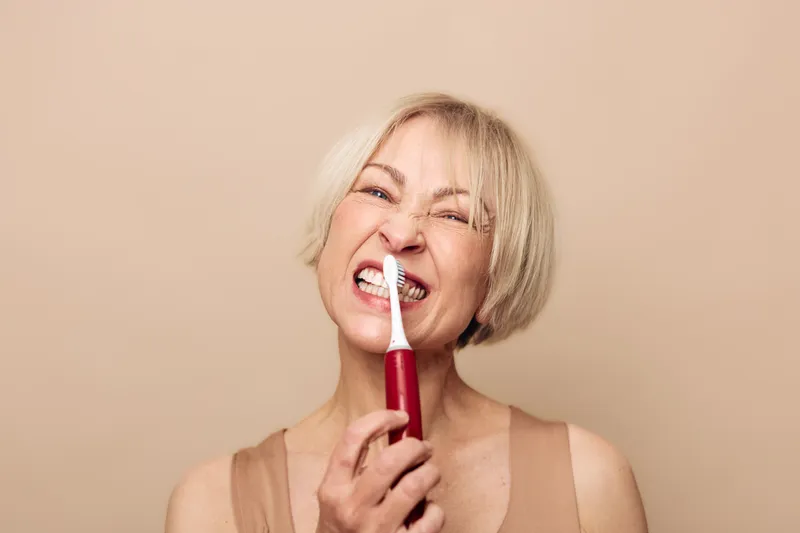 Middle aged woman using electric toothbrush with playful expression on beige background. Concept for dental care, oral hygiene, and healthy lifestyle for mature adults.