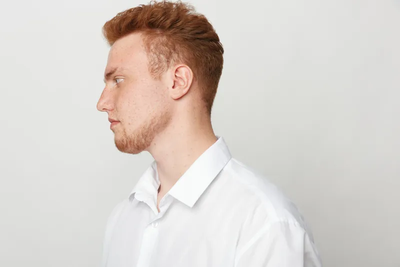 Profile of a young redhead man with beard wearing white shirt looking to the left on a plain light background. Concept of casual style, portrait, and thoughtful expression.