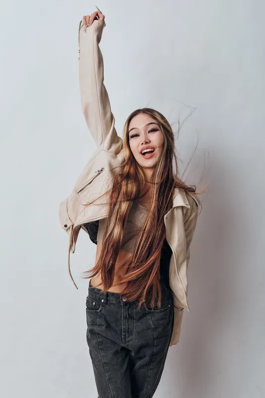 Young woman with long hair and braids wearing casual jacket and pants posing with raised arm and smiling on light background in studio. Joyful girl with confident expression.