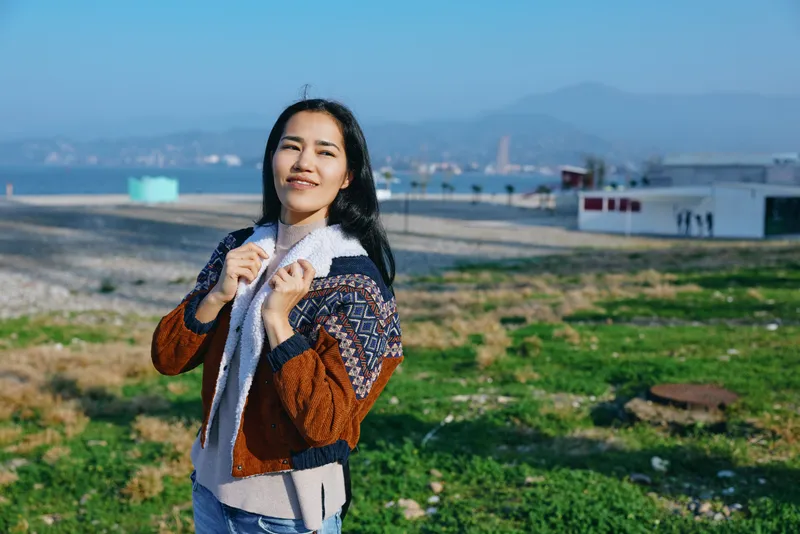 Woman outdoors wearing scarf and jacket by the ocean coast with distant mountains, bright sunlight, casual fashion, relaxed expression, natural landscape around, cheerful mood, travel lifestyle scene.