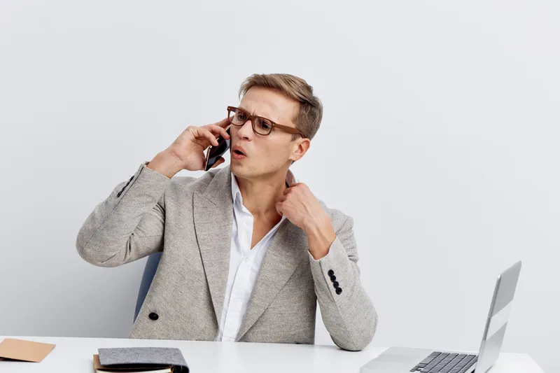 Portrait of a professional young caucasian man wearing glasses and a light gray suit, talking on a smartphone, sitting at a white desk with a laptop and notebooks, neutral background, business communi