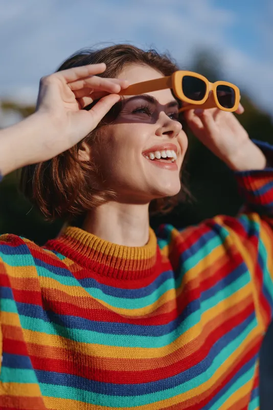 Young woman in a colorful striped sweater and yellow sunglasses, smiling while adjusting shades outdoors. Short brown hair, casual outfit, sunny travel vibe and joyful lifestyle portrait.