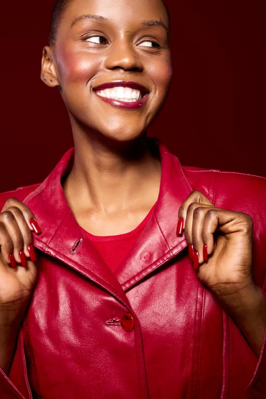 Smiling black woman with dark skin wearing a red leather jacket and matching nails poses confidently on red background. Fashion concept with vibrant colors and joyful mood.