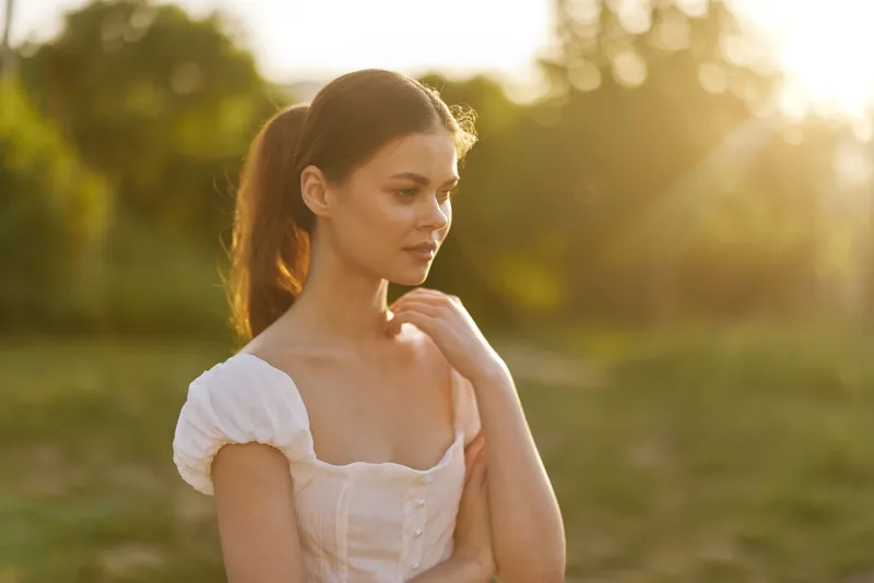 Young woman in a summer dress thoughtfully gazing into the distance during golden hour, surrounded by lush greenery and warm sunlight