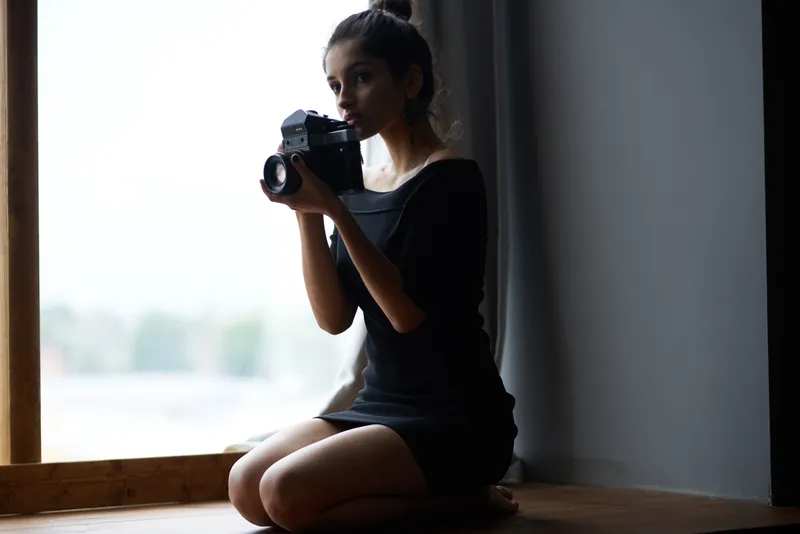 Красивая женщина держит фотоаппарат у окна, рядом декор, мода, стиль жизни, студия