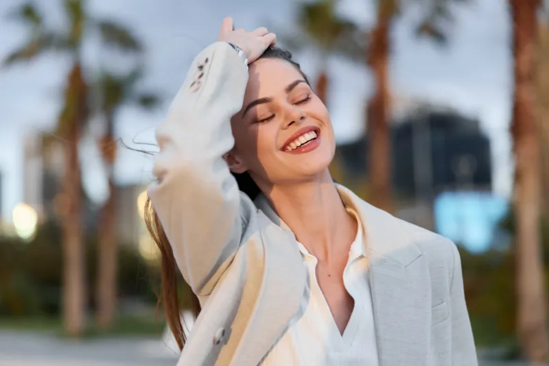 Confident businesswoman smiling outdoors in a stylish outfit, radiating positivity and strength amid a modern urban backdrop with palm trees.