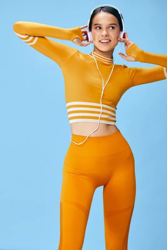 Woman fitness sport enthusiast in orange athletic wear, listening to headphones, posing against a bright blue background, capturing energy, balance and active lifestyle.