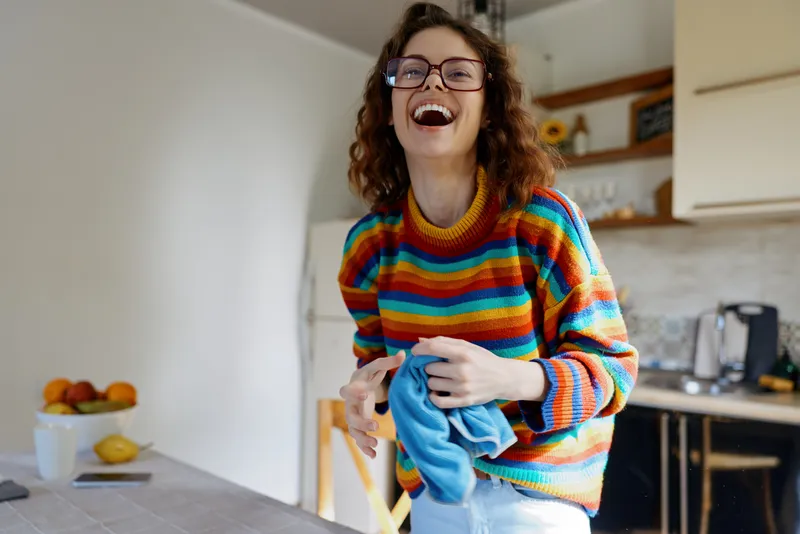 Joyful woman in a colorful striped sweater laughs in a bright kitchen while folding laundry, capturing a spontaneous home lifestyle moment and warm, candid everyday happiness