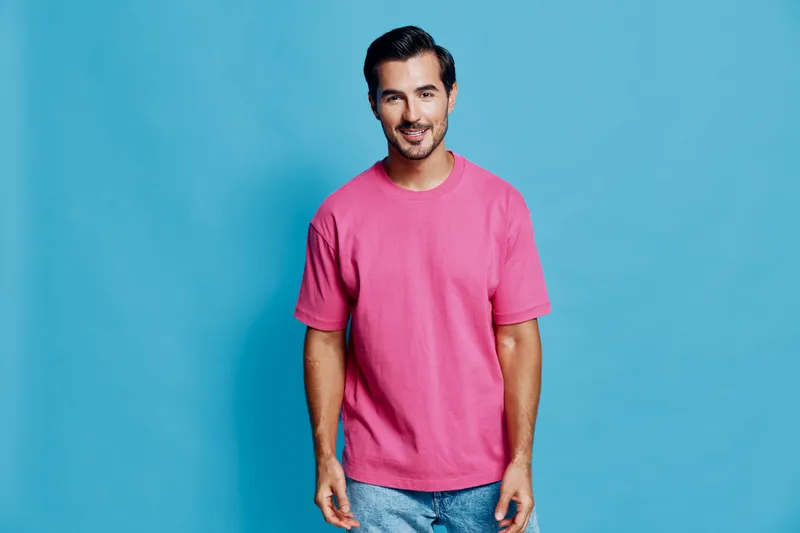 Full body smiling happy cheerful young man wearing a pink t-shirt stands against a bright blue background, showcasing a friendly and relaxed demeanor. People lifestyle concept