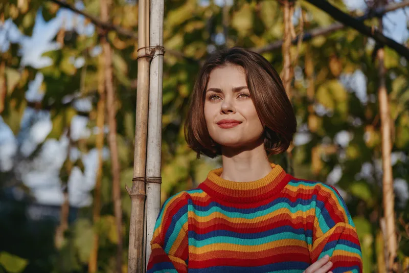 Woman in rainbow striped sweater, short brown hair, young adult smiling while standing in a backyard garden by bamboo trellis, casual outfit, relaxed lifestyle and outdoor portrait.