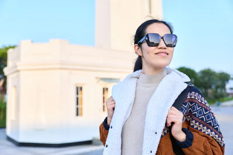 Stylish woman wearing sunglasses and a shearling jacket posing outdoors near a light building, showcasing a cozy knit sweater for travel fashion and everyday lifestyle