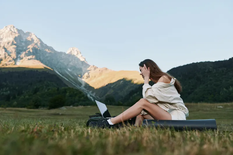 A relaxed woman sits on a mat in a wide meadow with a laptop, enjoying sunny weather and peaceful scenery, conveying focus, creativity, and a productive outdoor work mood.