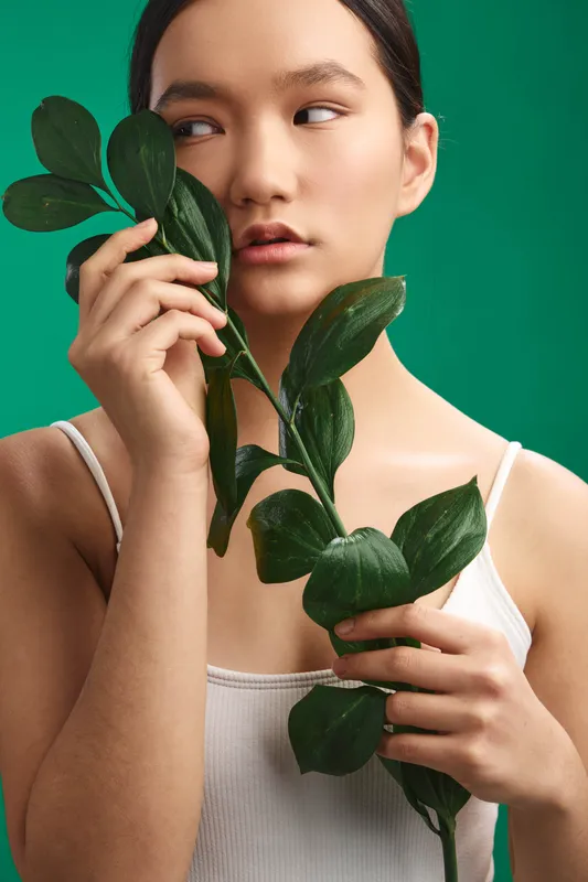 Asian woman holds a leafy stem close to her face in a studio portrait with a green background, conveying calm, natural beauty and a contemplative mood.