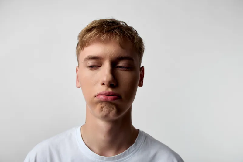 Close up of pouty face on a young man against a plain background, wearing a white t shirt, conveying casual mood, emotion and subtle humor in a minimal setting