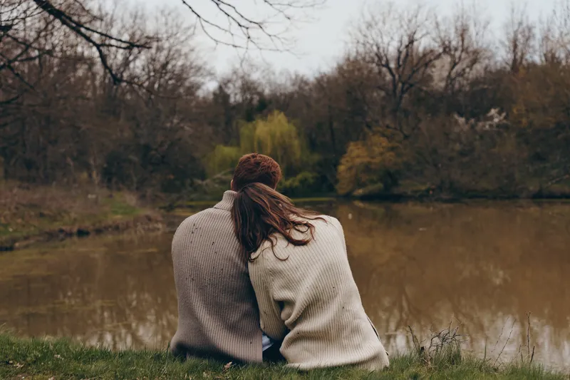 Two people sit close by a calm lake, warm sweaters embrace them as breeze rustles leaves, peaceful companionship in a natural, sunlit outdoor setting with muted greens and browns, capturing intimate,