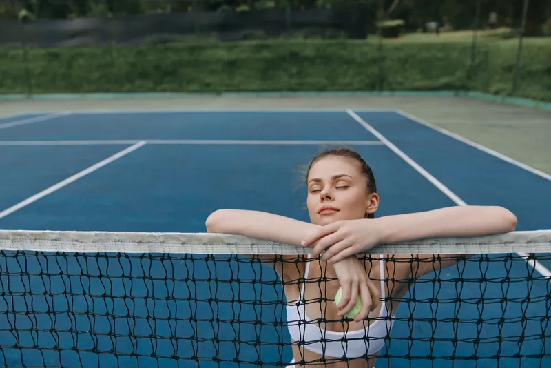 Young female tennis player relaxing at the court, wearing a sporty outfit, showcasing confidence and determination, with a vibrant blue background