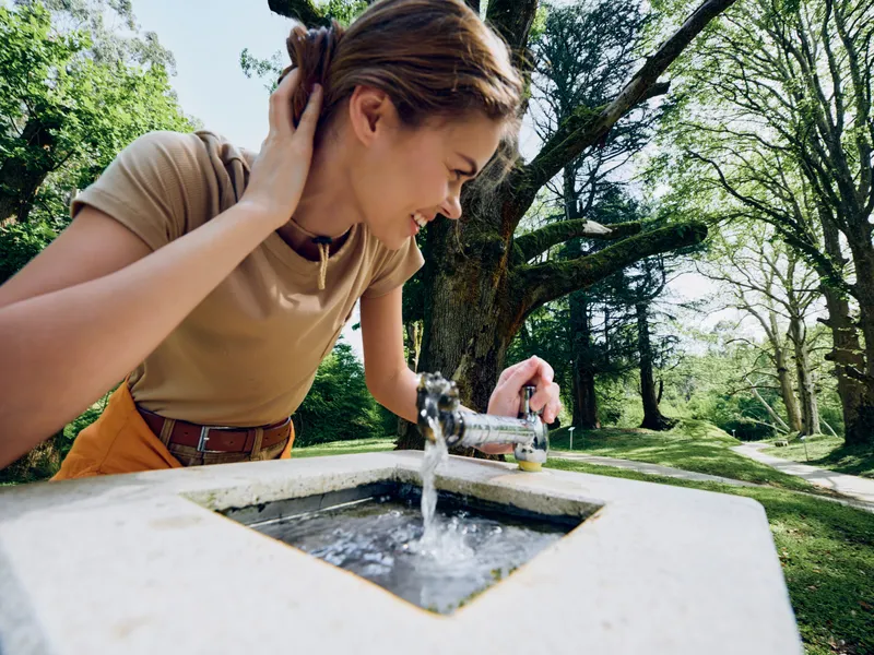 Woman drinks from a fountain in a park, bending to sip clean water from a metal spout, surrounded by trees and nature in warm sunlight, casual summer moment of refreshment.