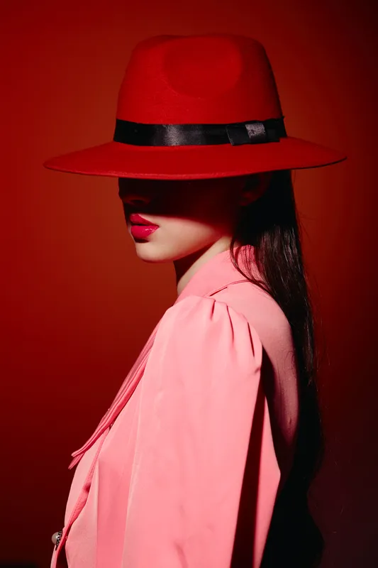 Fashionable woman with long dark hair wears vibrant red hat and pink outfit, portrait in low shadow on red background. Creative style photo for beauty and fashion themes.