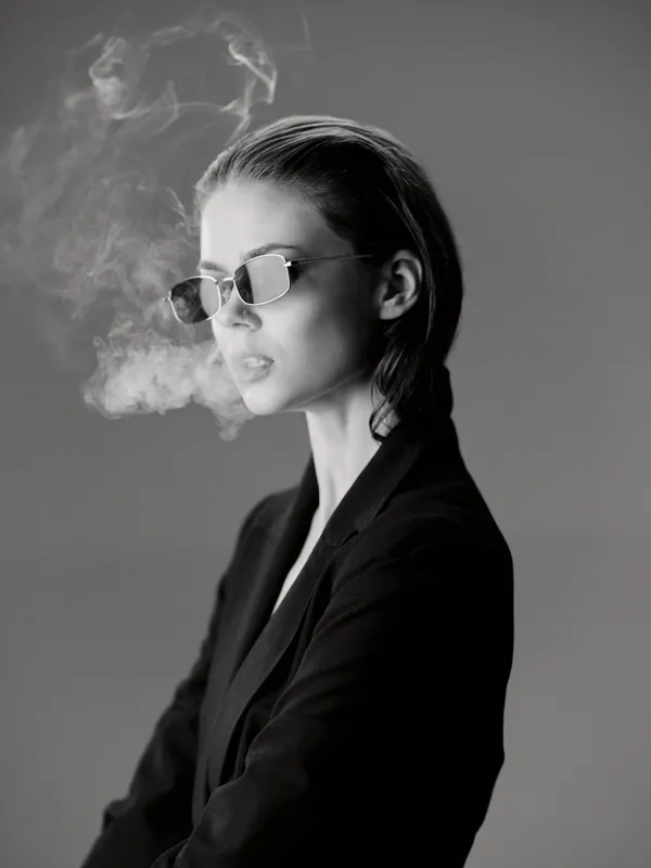 Black and white portrait of a stylish woman wearing sunglasses exhaling smoke in a minimalistic studio setting.
