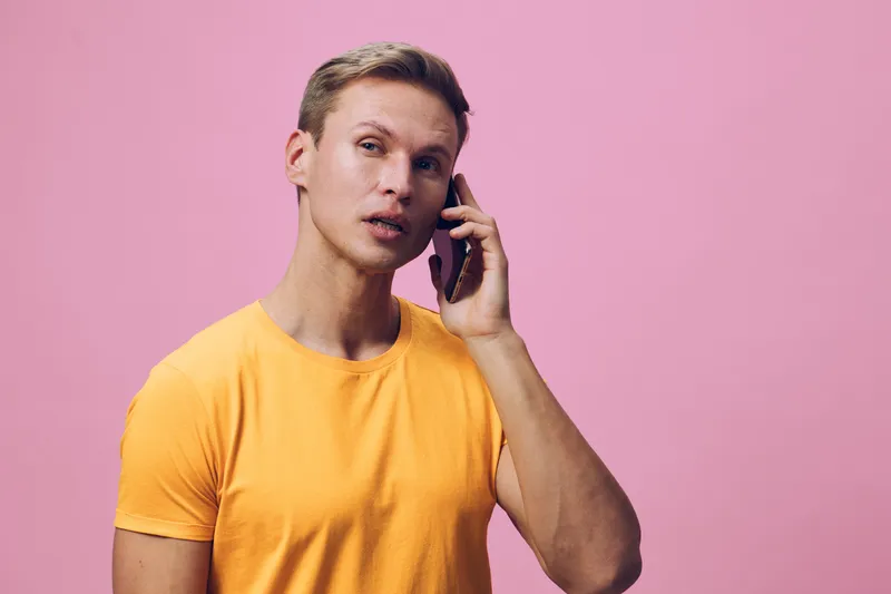 Smiling man with short light brown hair wearing yellow t-shirt talking on mobile phone against pink background. Studio portrait of a cheerful young male in casual clothing, natural expression, isolate
