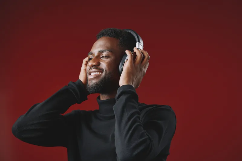 Young African man enjoying music with headphones on a vibrant red background, expressing joy and relaxation in a stylish black outfit