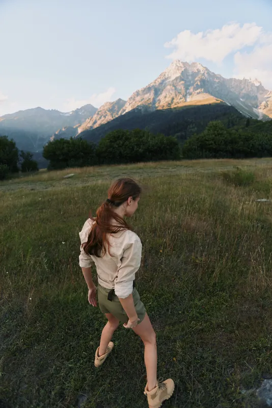 Young woman with brunette hair walks through a grassy field toward distant mountains, expressing curiosity and calm in a sunlit outdoor moment, her light beige shirt and olive shorts fluttering in a